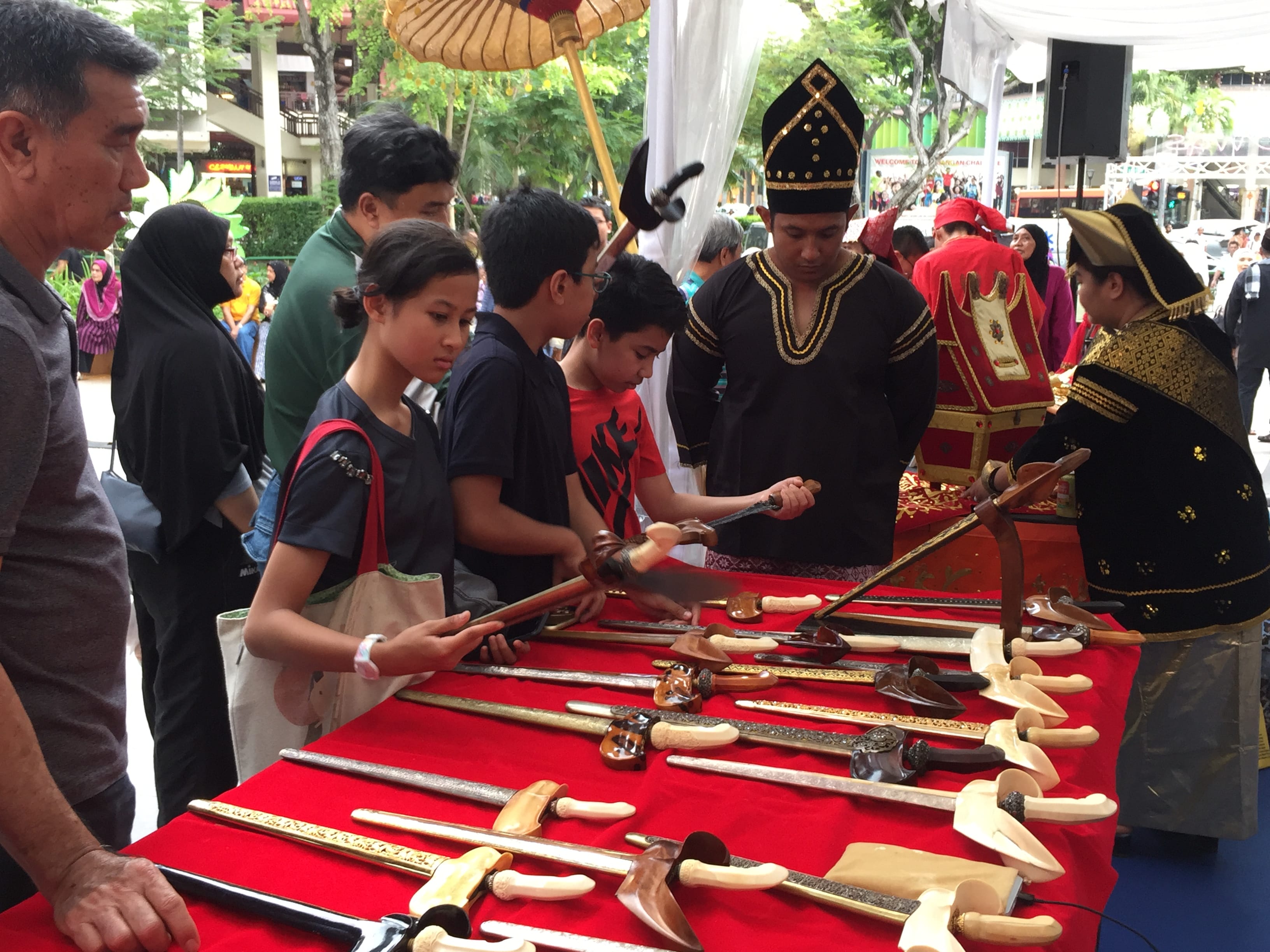 People in traditional costumes and visitors examine a display of ornate bladed weapons on a red cloth table at an outdoor cultural event.